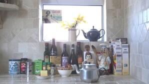 Un comptoir de cuisine sur lequel on trouve divers objets, notamment une théière, des bouteilles, un bol, et une fenêtre donnant sur des fleurs jaunes.