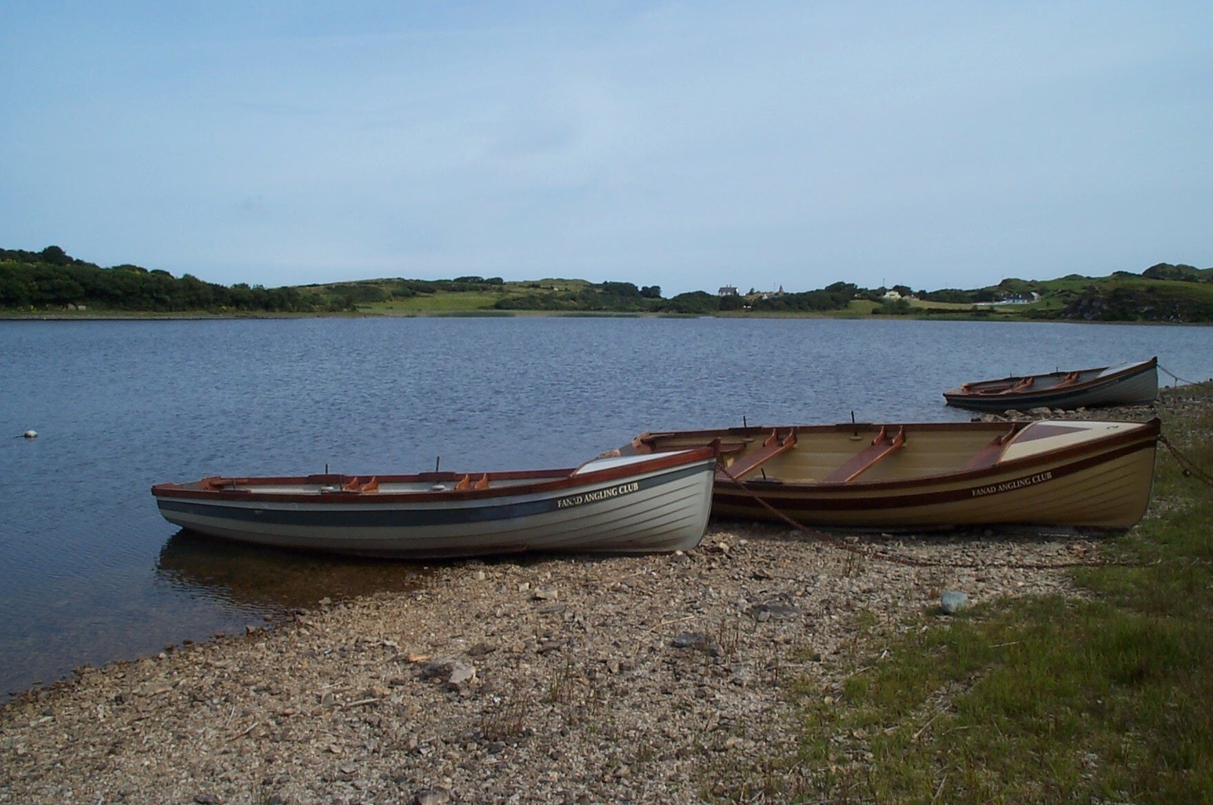 Three rowboats named "Fanad Angling Club" docked on a pebbly shore by a calm lake with a grassy and hilly landscape in the background.