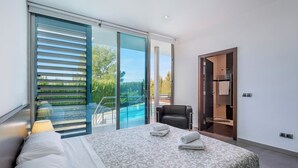 Ein modernes Schlafzimmer mit einem großen Bett, einem Sessel, Blick auf einen Swimmingpool und einer Schiebetür zu einem Außenbereich.