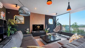 Ein modernes Wohnzimmer mit großem Flachbildfernseher, einer Couch, einem Couchtisch und einem Hängekorbstuhl. Ein großes Fenster bietet einen Blick ins Freie.
