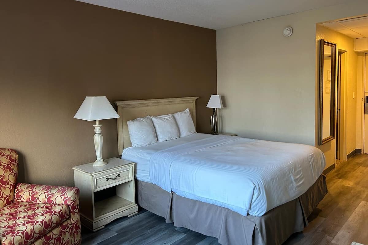 Habitación de hotel con una cama, dos mesitas de noche con lámparas, una silla y una puerta que da a otra habitación.