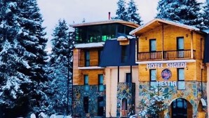 Ein zweistöckiges Holzgebäude mit Balkon, mit der Aufschrift "HOTEL SHGEDI", umgeben von schneebedeckten Bäumen.
