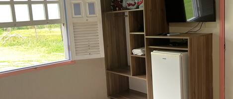 A room with a bed, a small white refrigerator, a wooden wardrobe, a television mounted on the wall, and a window with a view of greenery.