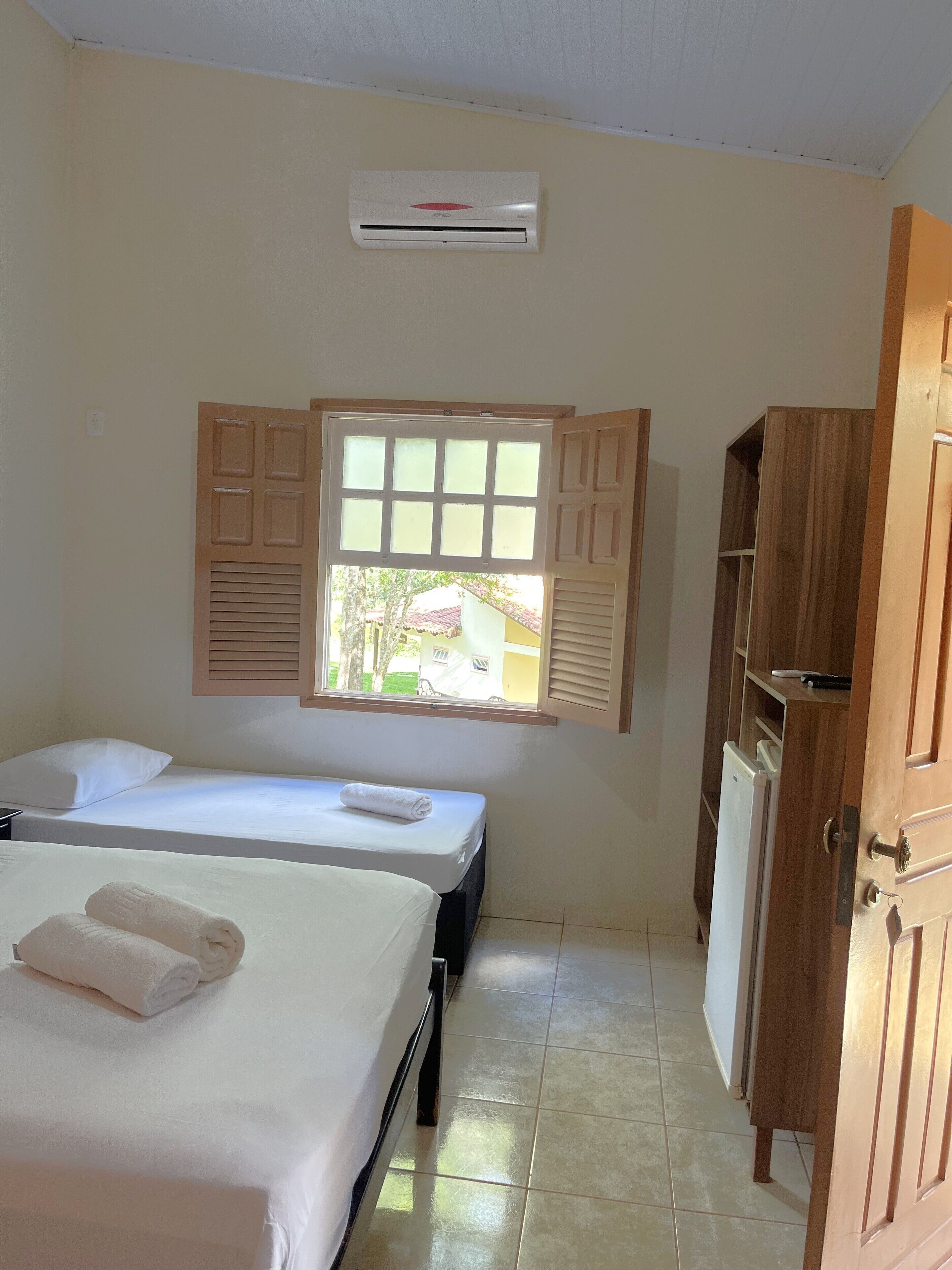 A hotel room with two beds, a window with shutters, an air conditioning unit, and a wooden wardrobe.