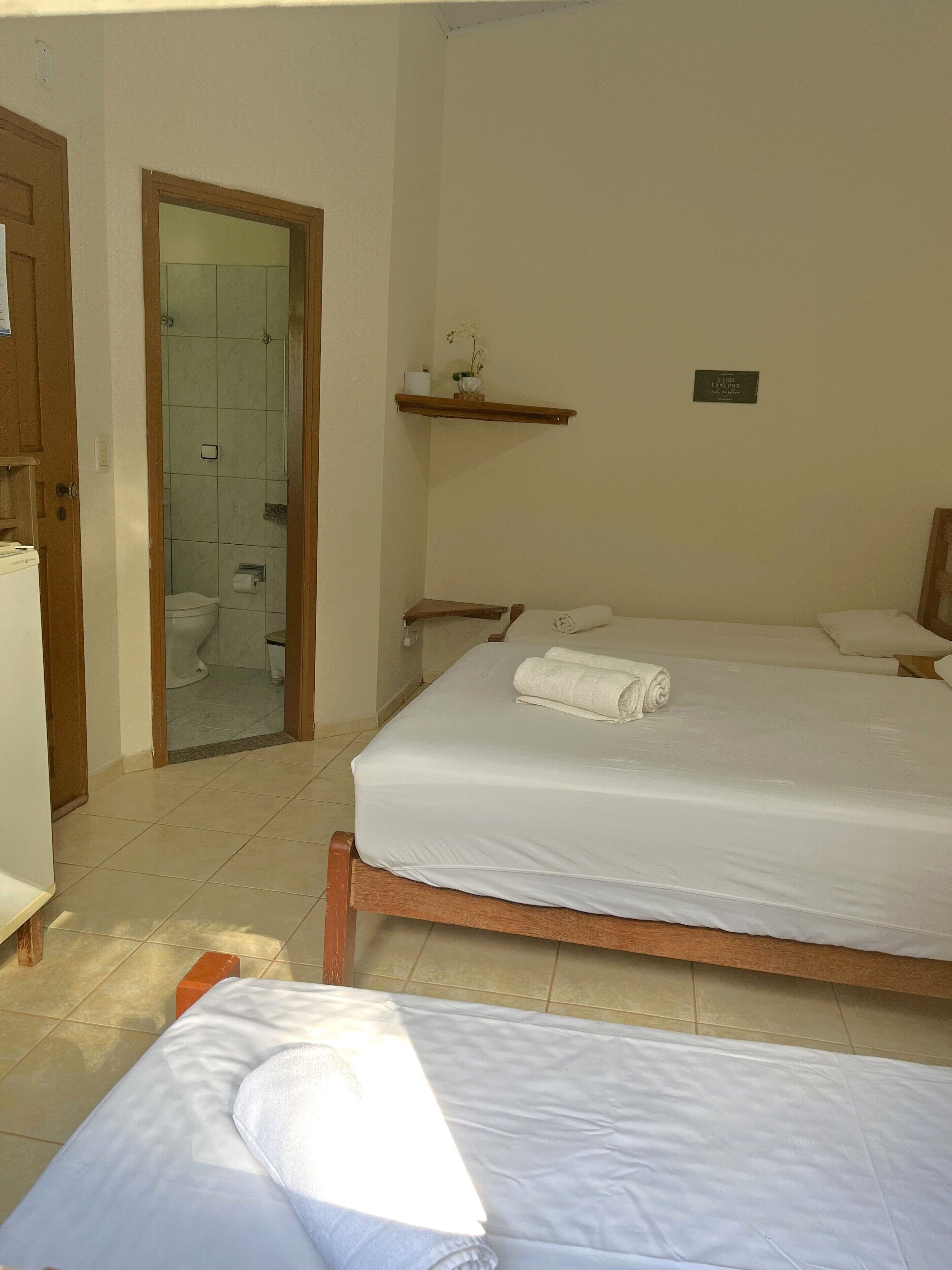 Quarto de hotel com duas camas, cabeceira de madeira, um pequeno aparador com um vaso e um espelho na parede.