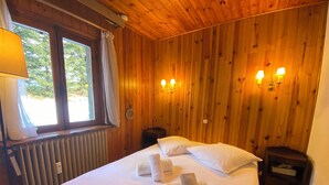 Une pièce aux murs lambrissés, avec un lit, deux lampes de chevet, une fenêtre avec des rideaux et un radiateur.