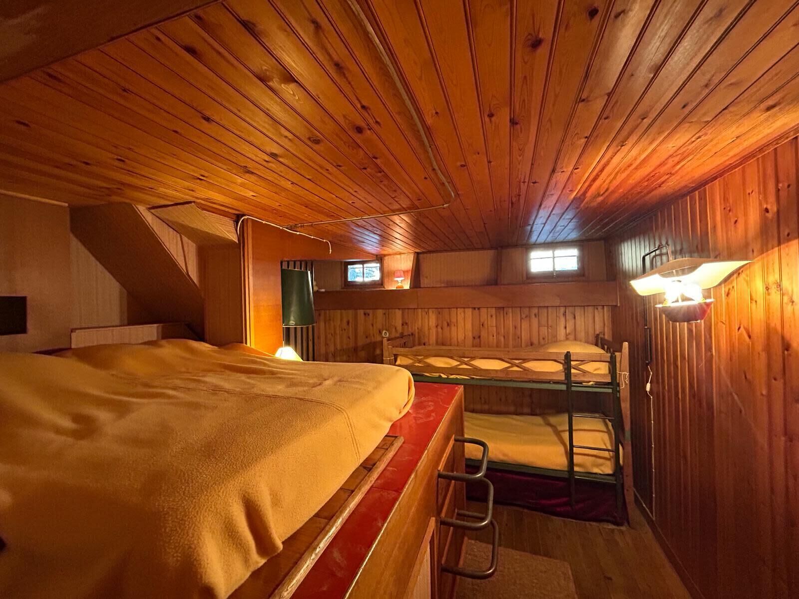 A cozy attic bedroom with a bunk bed and a single bed, wooden walls, and a sloped ceiling.