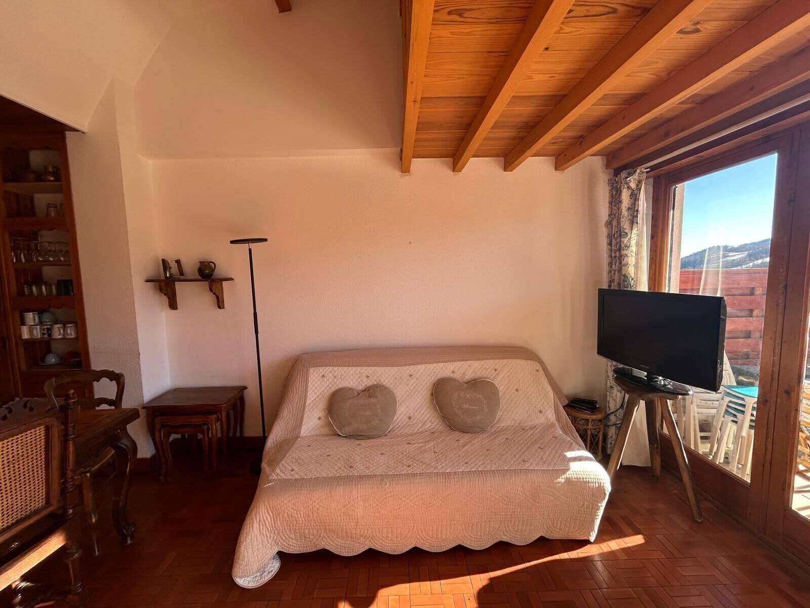 Una habitación acogedora con techo de madera, un sofá con cojines con forma de corazón, un televisor sobre un mueble bajo y una ventana con vistas a edificios.