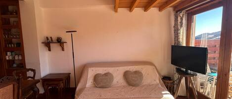Una habitación acogedora con techo de madera, un sofá con cojines con forma de corazón, un televisor sobre un mueble bajo y una ventana con vistas a edificios.