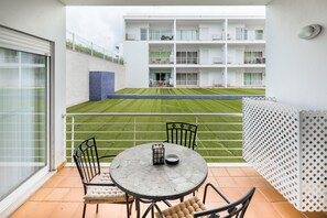 A balcony with a round table and chairs, a view of a green lawn, and a multi-story building in the background. - Fuseta Formosa Village (Olhao)