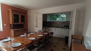 Un comedor con una mesa de madera dispuesta para seis personas, rodeada de sillas. Hay una zona de cocina con un microondas y una ventana sobre el fregadero.