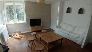 Una sala de estar con una mesa de centro de madera, cuatro taburetes de madera, un sofá blanco, un televisor sobre un mueble de madera y una ventana con vistas a árboles.