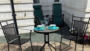 Une terrasse avec une table ronde et quatre chaises, un petit jardin agrémenté de fleurs et, au fond, un bâtiment blanc.