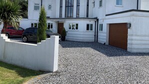 Une maison moderne à deux étages, avec une allée gravillonnée, une voiture rouge et une voiture noire garées dans l’allée.