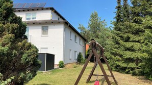 Un ensemble de balançoires en bois installé dans une cour verdoyante, avec une maison en arrière-plan.