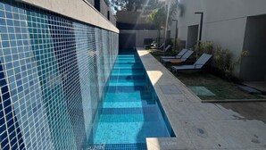 Una piscina con borde de azulejos, rodeada de sillas de descanso y un pequeño jardín.