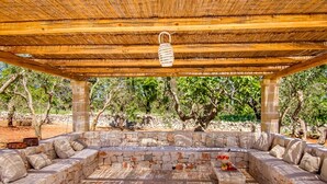 Un espace extérieur aménagé avec des sièges, abrité et doté de murs en pierre et de coussins, le tout entouré d’arbres.