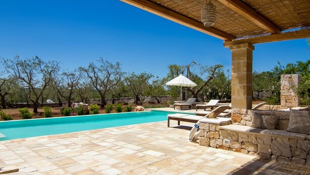 Un espace aménagé au bord de la piscine, doté d’un sol en pierre, de chaises longues, d’un coin salon abrité et offrant une vue sur les arbres et un ciel dégagé.