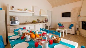 Une cuisine moderne avec des armoires blanches, un four à micro-ondes et une cuisinière. On y trouve une table à manger avec des chaises bleues, des assiettes et divers aliments. Un téléviseur est fixé au mur, et un canapé est visible au fond de la pièce.