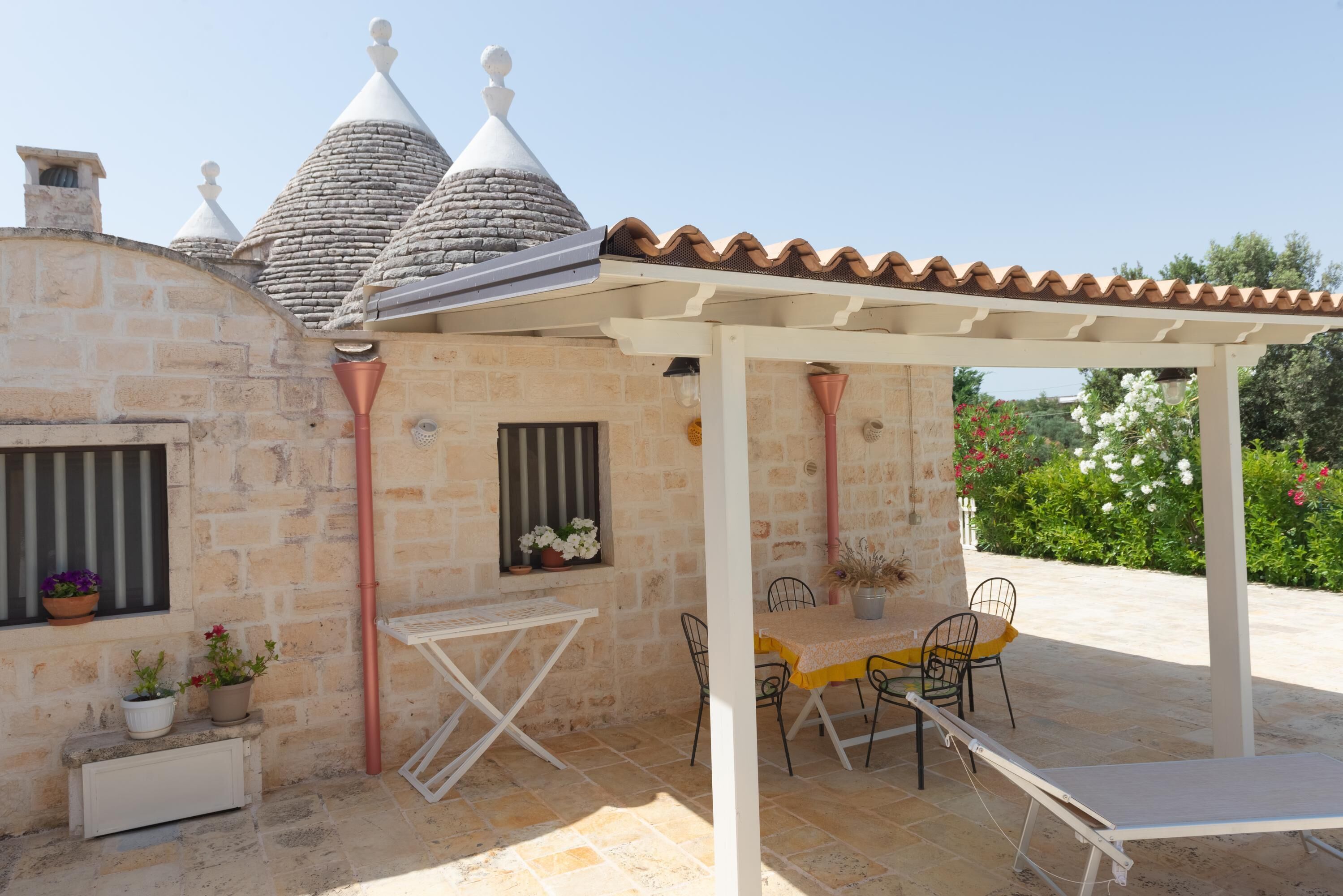 A stone building with a tiled roof, a covered patio with a table and chairs, and a window with potted plants.