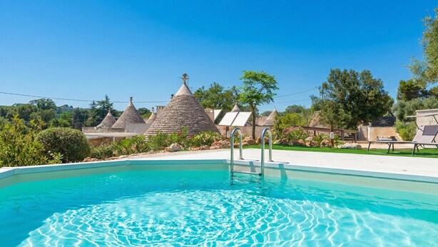 A swimming pool with clear water, surrounded by stone-roofed buildings and lush greenery. - Trulli Dei Ciclamini - Locorotondo (Locorotondo)