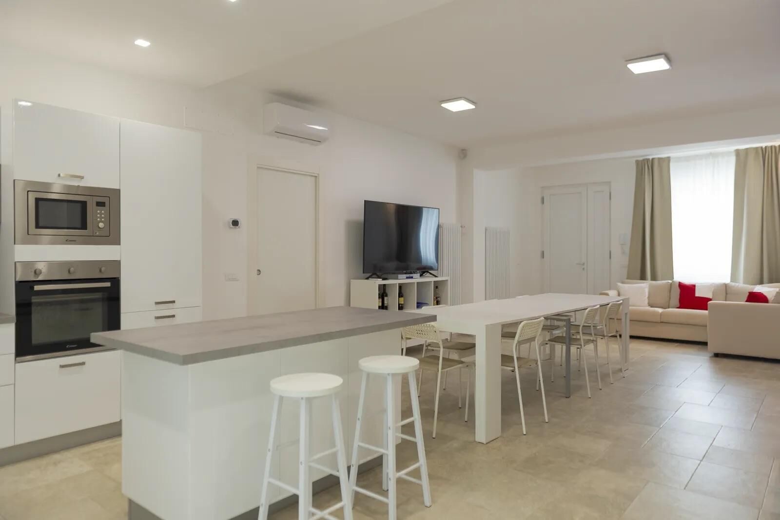 Een moderne keuken met een centraal eiland, barstoelen, een eetgedeelte met stoelen, een flatscreen televisie en een zithoek met een bank en gordijnen.