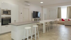 A modern kitchen with a central island, bar stools, a dining area with chairs, a flat-screen TV, and a living area with a sofa and curtains.