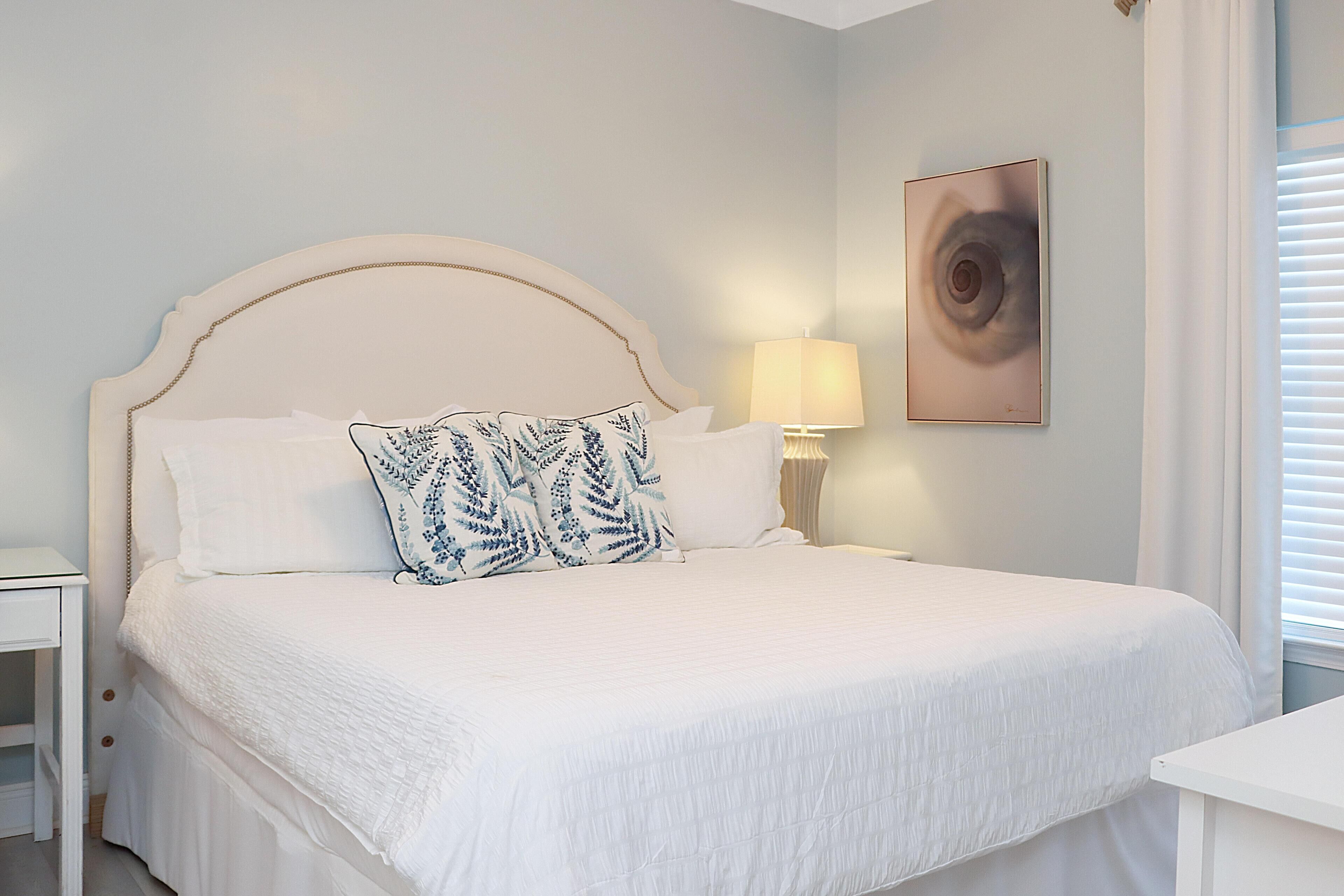 A neatly made bed with a white headboard and a patterned pillow, a bedside lamp, and a framed artwork on the wall.