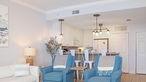 A living room with a white sofa, two blue armchairs, a coffee table, and a lamp. There is a dining area with a table and chairs in the background. - Summerhouse 309 (Port St. Joe)