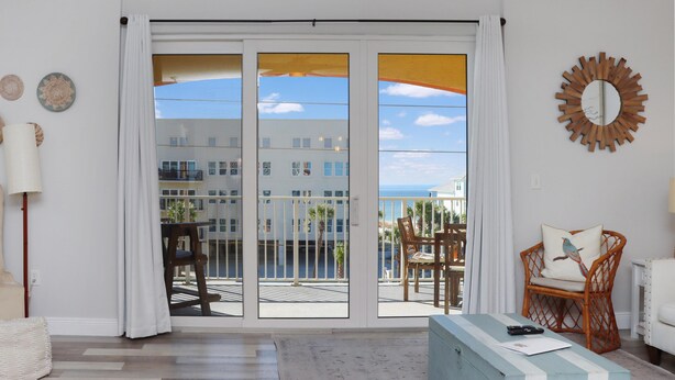 A room with a large sliding glass door leading to a balcony with a view of buildings and the sea. There is a wicker chair and a white armchair on the balcony, and a wooden side table with a lamp. - Summerhouse 309 (Port St. Joe)