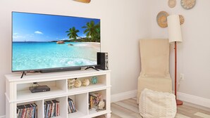 A living room with a flat-screen TV on a white stand, a bookshelf with various items, a beige armchair, and a floor lamp. - Summerhouse 309 (Port St. Joe)