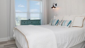 A bedroom with a large bed, a nightstand, a window with blinds, and a wall-mounted lamp. - Summerhouse 309 (Port St. Joe)