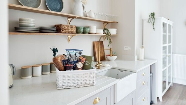 A kitchen with white cabinets, a marble countertop, and shelves holding plates, bowls, and a basket of groceries.