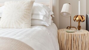 A neatly made bed with white linens and a textured pillow, a round wooden side table with a lamp and a candle, and a wall-mounted decorative piece.