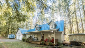 A two-story cabin with a blue roof, a porch with red chairs, and a small shed to the left. - Relaxing River Lodge • Hot Tub • Family Friendly (Rhododendron)