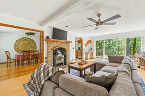 A living room with a fireplace, a sofa, a dining area, and a large window with a view of trees. - Relaxing River Lodge • Hot Tub • Family Friendly (Rhododendron)