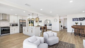 A modern kitchen with white cabinetry, stainless steel appliances, and a central island. There are two white armchairs and a bar area with stools.