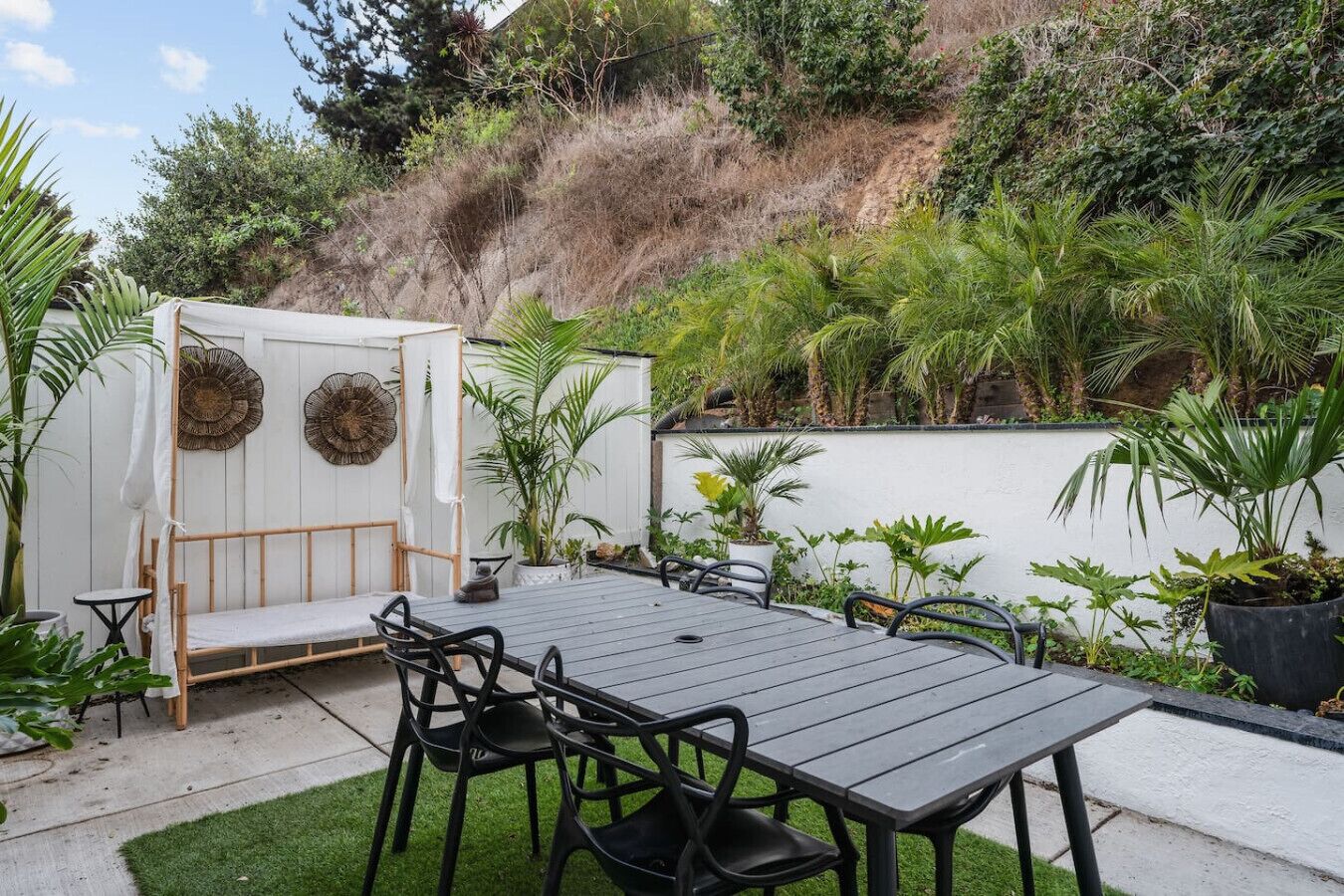 A patio with a wooden table and chairs, surrounded by potted plants and a white wall.