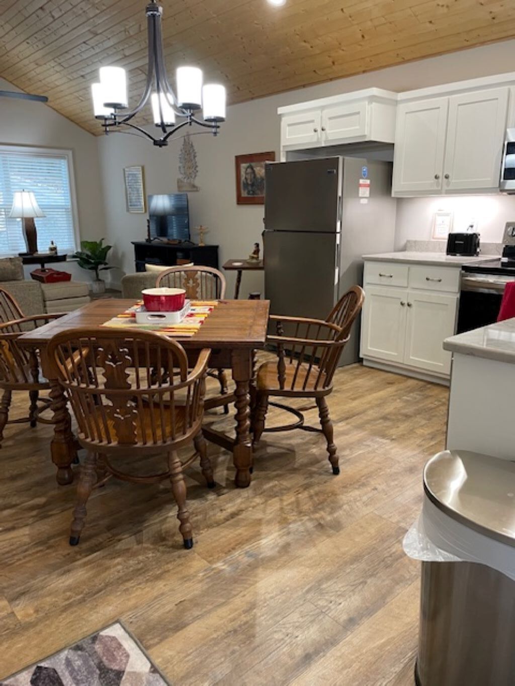 A dining area with a wooden table and chairs, a chandelier, and a kitchen with white cabinets and stainless steel appliances.