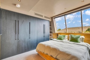 A bedroom with a large wardrobe, a bed with green pillows, and a view of the outside through a large window. - Kings House (London)