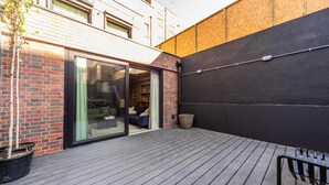 Une terrasse extérieure moderne avec une plateforme en bois, une porte coulissante en verre donnant sur un espace intérieur et un mur noir.