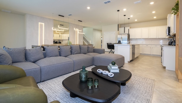 Un salon moderne avec un canapé d’angle gris, une table basse sombre agrémentée d’objets décoratifs, et une cuisine en arrière-plan avec des meubles blancs et un réfrigérateur en acier inoxydable.