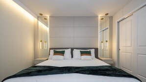 Un lit bien fait, avec du linge de lit blanc et deux coussins verts et bruns, une couverture vert foncé et une tête de lit agrémentée d’un miroir.