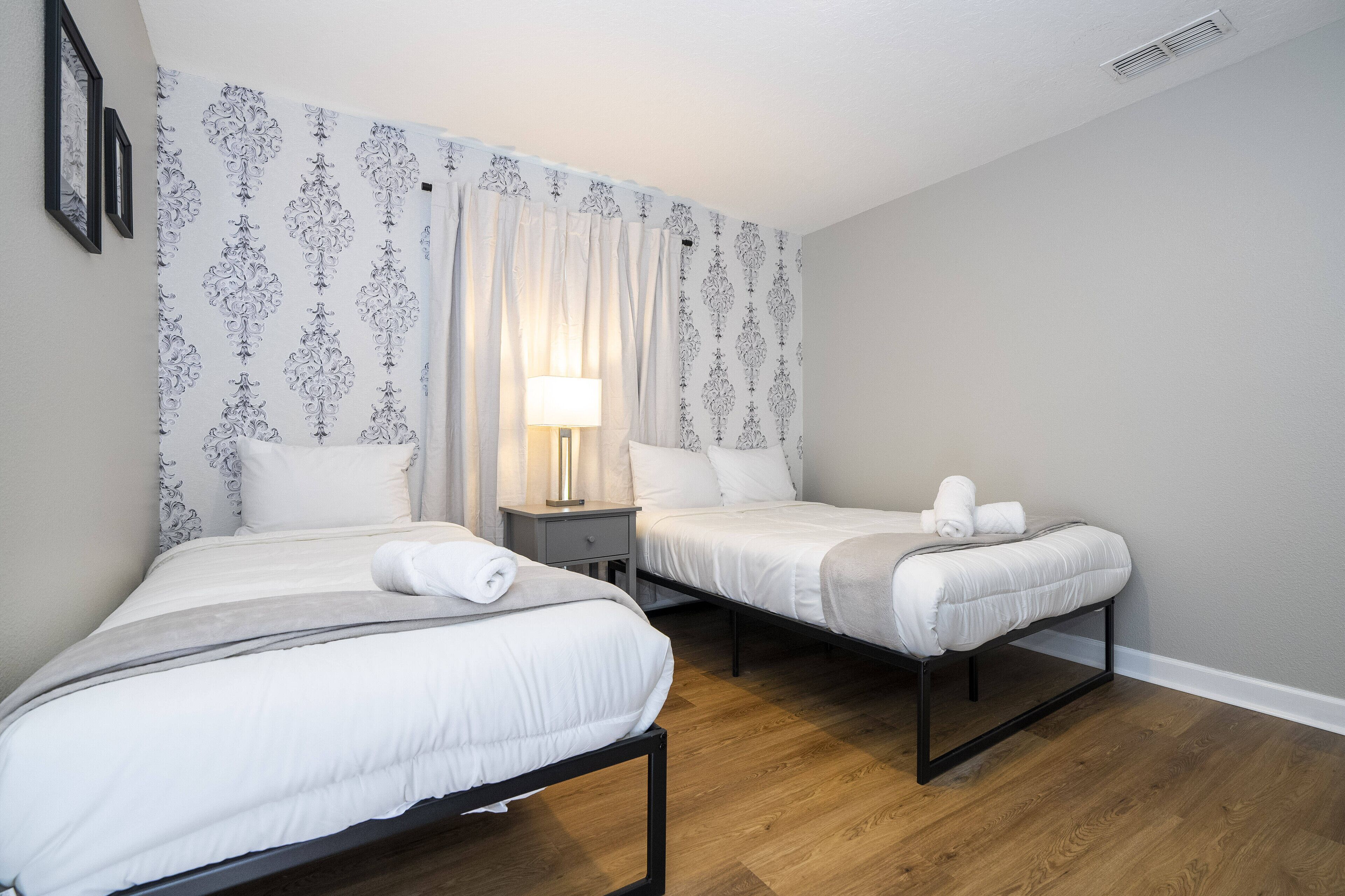 A hotel room with two single beds, a nightstand with a lamp, and a patterned wallpaper.