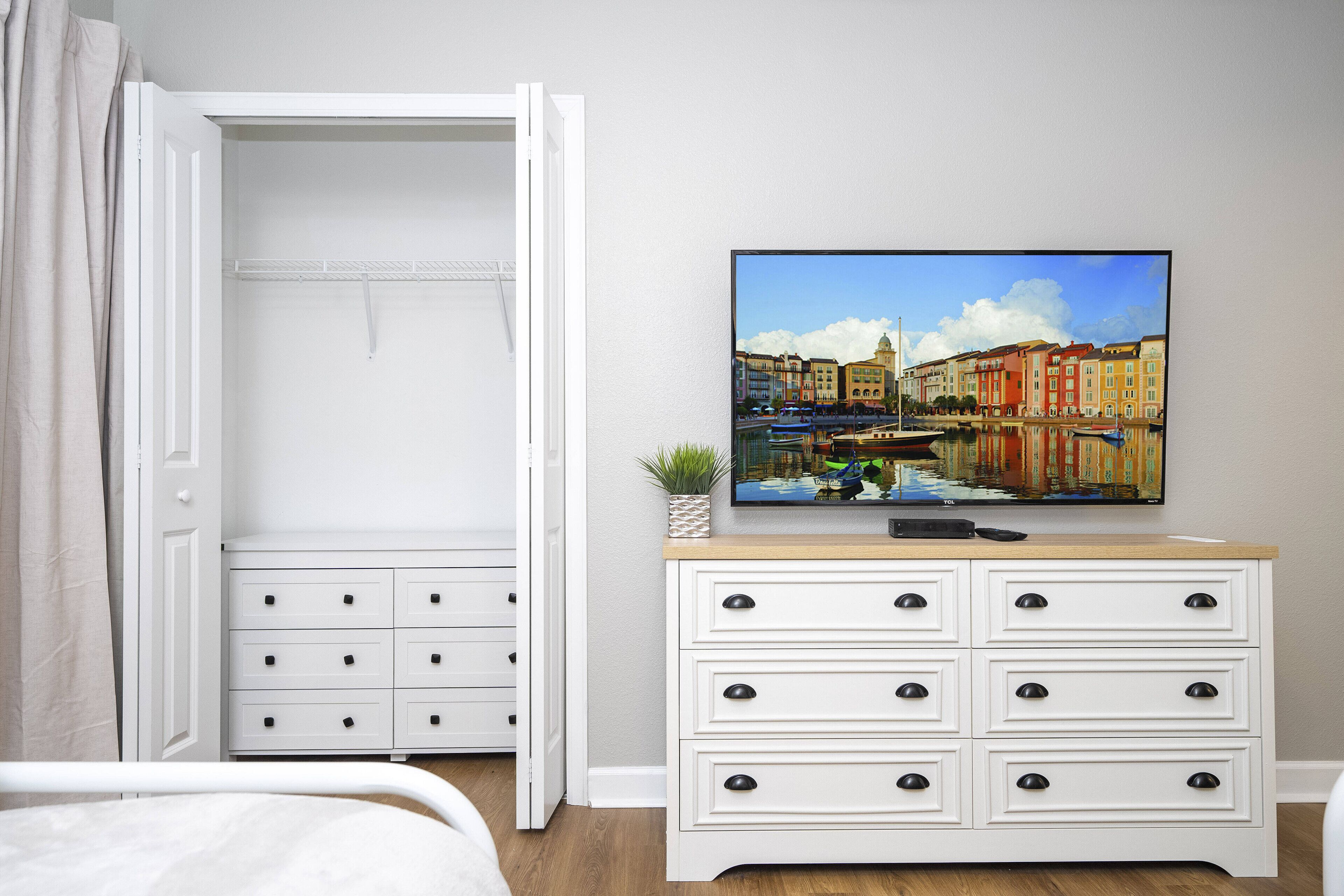 A bedroom with a bed, a white dresser with drawers, a television on top, and a closet with a door open.