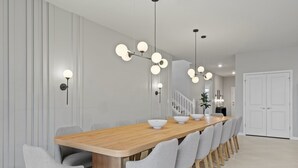 Un espace de salle à manger moderne, avec une longue table en bois, des chaises à revêtement gris et des luminaires suspendus dotés de plusieurs ampoules sphériques.