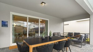 Une terrasse extérieure avec une table en bois et des chaises noires, un coin salon avec des canapés et une table basse, et une porte vitrée donnant sur un espace intérieur.