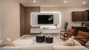 A modern living room with a sofa, two side tables, a flat-screen TV, a kitchen area with cabinets, and a countertop.
