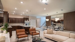A spacious living area with a sofa, armchairs, and a coffee table. There is a kitchen in the background with wooden cabinets and a dining area with a table and chairs.