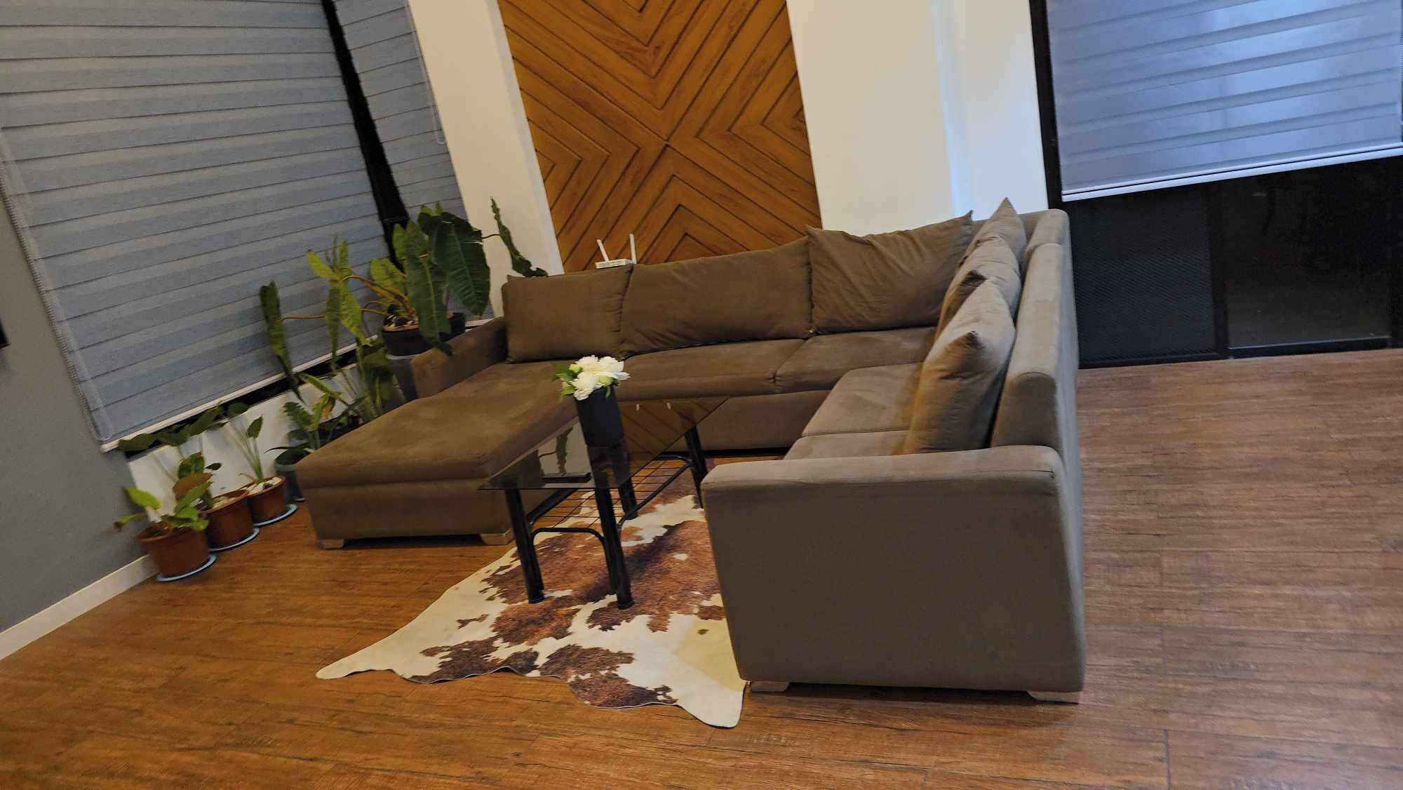 Una sala moderna con un sofá modular, una mesa de centro de vidrio y un tapete de cuero de vaca. Hay plantas en macetas y una pared de acento de madera.